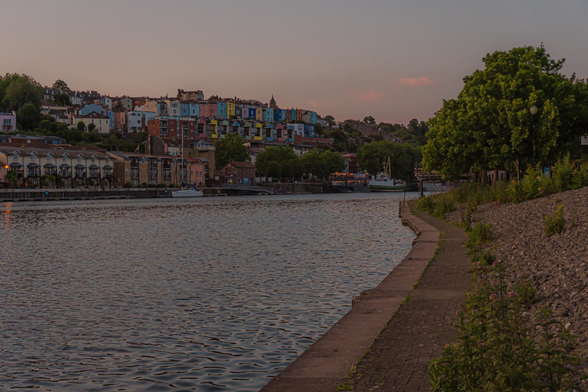 On the right is a path along the river. On the left is the river and left top is a series of colourful terraced houses. It is around sunset and the whole picture has a pinkish hue.