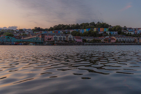 The foreground is almost level with the water and looking across it you see the colourful terraced houses and a turquoise Bridge. It is around sunset and there is a peachy hue that is also reflected in the water.
