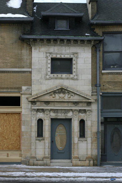 A color photo of a row of stone and brick buildings. Centered in the frame is a white stone building with a somewhat ornate facade and a black roof. Above the door, the stone is carved to read "GERHART BLOCK". The street and sidewalk in front of the building is snowy.