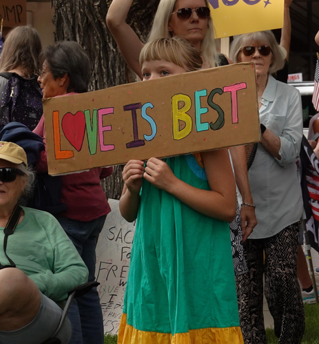 a young girl in a green dress holds a hand written and colored sign on cardboard which says "love is Best". She holds it in front of her face, so that only her eyes and blonde bangs show