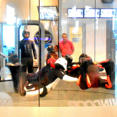 The image depicts an indoor wind tunnel facility. In the foreground, several individuals are suspended in mid-air, wearing full-body flight suits and helmets, simulating the experience of flying. The suits are predominantly black with red accents, and the helmets are black and red. One person is wearing a white helmet. The individuals appear to be in various stages of flight, with some leaning forward and others extending their arms.

In the background, a man in a red striped shirt stands near a glass door labeled "SALA DE VUELO," which translates to "Flight Room" in Spanish. The door has a circular logo with a stylized "P" on it. The facility has a modern design with glass walls and a circular floor, allowing for a 360-degree view of the activity. The lighting is bright, and the environment appears clean and well-maintained. The image captures the dynamic and exhilarating nature of indoor wind tunnel flying.

Provided by @altbot, generated privately and locally using Ovis2-8B

🌱 Energy used: 0.211 Wh