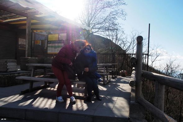Farbfoto, gegen die Sonne aufgenommen. Zwei Frauen sitzen in Bildmitte auf einer Picknickbank aus Holz. Das Haar der Frau links wird sichtlich vom Wind verweht, der so stark bläst, dass der geplante Besuch des Vesuvs an diesem Tag im Januar abgebrochen werden musste.