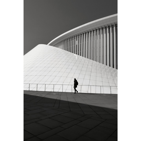Black and white street photography taken in Luxembourg city. Somebody passes by the Philharmonie building whose whiteness contrasts with the dark space created by the shadow cast from the tower behind the photographer.