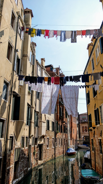 Szene in Venedig. Zwei Häuserzeilen an einem Kanal zwischen denen mehrere Wäscheleinen gespannt sind. An zwei leicht wehenden Bettlaken erkennt man, dass ein bisschen Wind geht. Die linke Häuserzeile spiegelt sich im Wasser des Kanals. Der Himmel ist ruhig und blau.