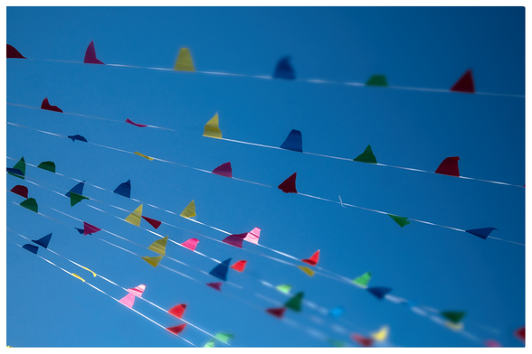 Blick in den blauen Himmel, viele kleine bunte Wimpel in gelb, blau, grün und rot sind an mehreren Schnüren befestigt, die von links oben nach rechts unten gespannt sind. 