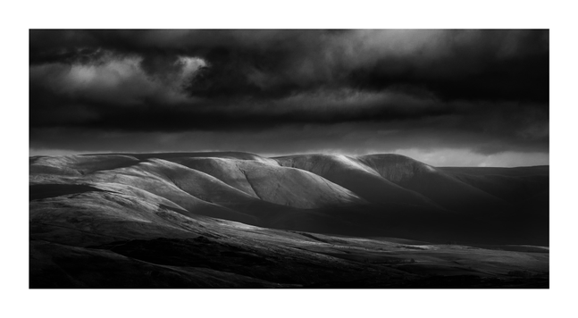 A black-and-white landscape photograph in wide panoramic format, framed with a white border. The image depicts a range of softly contoured hills under a dramatic sky. Patches of light break through heavy, textured cloud cover, illuminating sections of the undulating terrain and creating high contrast between sunlit slopes and shadowed valleys. The hills appear smooth and almost sculptural, with no visible vegetation or structures, emphasizing form and tonal gradation. The overall mood is tense and atmospheric, shaped by the interplay of light, shadow, and impending weather.