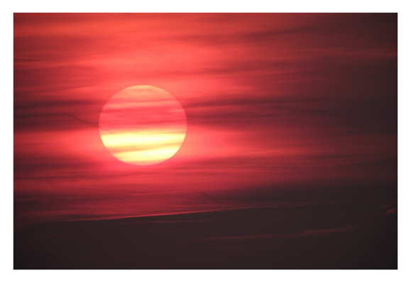 Foto im Querformat. Teleaufnahme der untergehenden Sonne. Der gelbe runde Kreis der Sonne ist umgebe von orange-roten und schwarzen Schlieren der Wolken und des Himmels. Es sieht aus wie ein Gemälde.