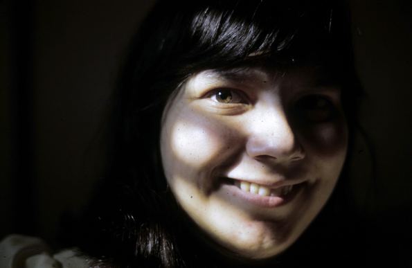 Photo of a dark haired young woman taken in natural light. Widow blinds cast shadows arcoss her fave. She smiles gently toward the camers. The background id black.