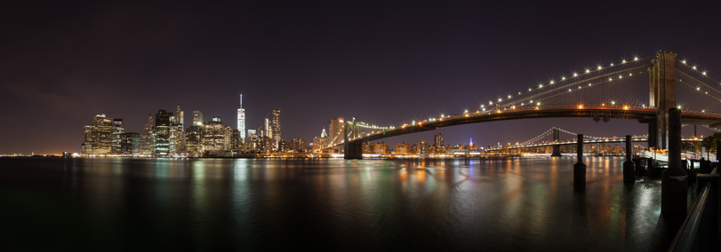 Panoramic night view of the New York City skyline with the Brooklyn Bridge and illuminated skyscrapers reflecting on the water's surface.