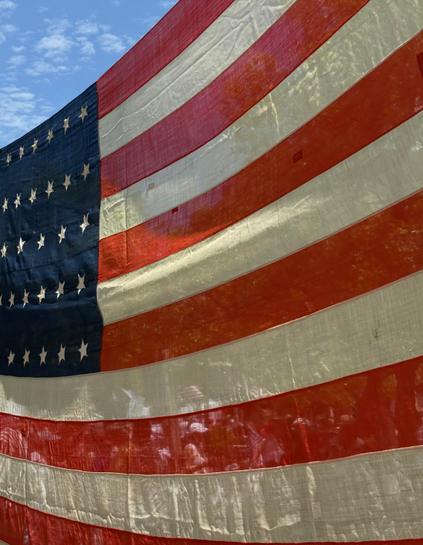 Photo of a large (roughly 25 by 15 feet) worn American flag with 35 stars that almost complete fills the image. It is being held up across an area where people are walking - we can see them through the thin red and white stripes of the flags. In the top left corner above the field of stars is a small triangle of blue sky with a few wispy clouds. 