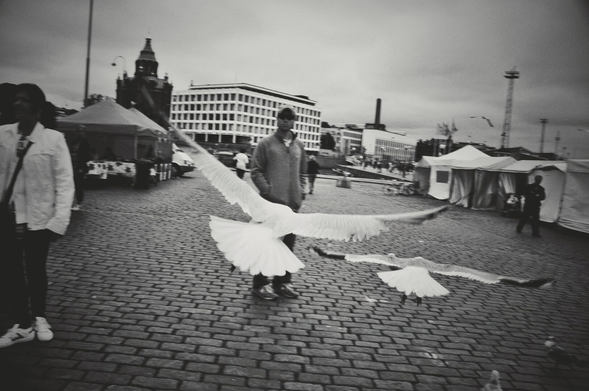 This is a landscape-orientated black and white, eye-level, medium shot of a cobblestone square where two large birds are captured mid-flight, seemingly taking off or landing. In the foreground, a person stands on the left, partially cropped out, wearing a light-colored jacket and dark pants, with white sneakers. In the middle ground, another person, wearing a dark cap and a light-colored jacket over dark pants, stands facing the camera. Behind them, several large tents are set up, and in the background, multi-story buildings and a church steeple are visible under an overcast sky. Other indistinct figures are present in the distance.