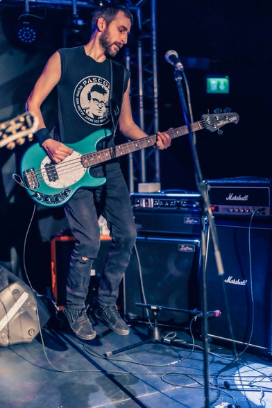 A musician playing an electric bass guitar while jumping on stage, wearing a sleeveless black shirt and dark pants. The setting features amplifiers in the background and stage lighting, creating a vibrant concert atmosphere.