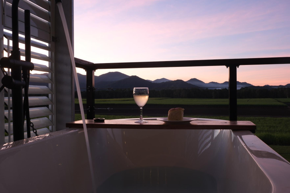 Photo of an outdoor bath tub at dusk, filling with water. There is a board across the bath with a wine glass and bun on it. In the background are some mountains.