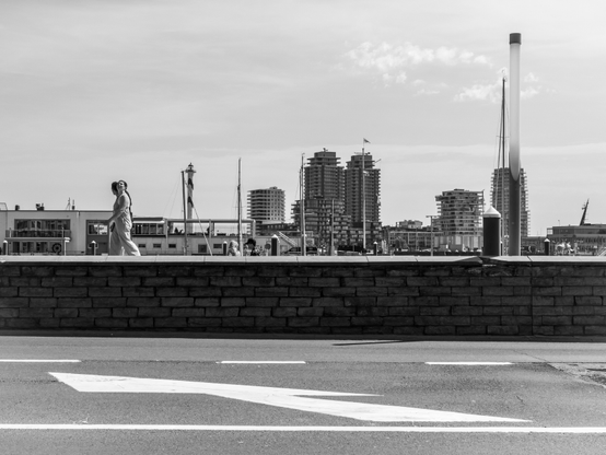 The image is a black and white photograph depicting a cityscape with a waterfront. In the foreground, a low stone wall runs horizontally across the image, with a pedestrian walking along it. The pedestrian is wearing a light-colored dress and appears to be moving towards the right side of the frame. The road in the foreground has a white arrow painted on it, indicating the direction of traffic.

In the background, a variety of buildings are visible, including high-rise residential structures and a prominent tall, slender tower with a flag at the top. Several masts, likely from boats, are visible, suggesting a marina or dock area. The sky is partly cloudy, and the overall atmosphere is calm and serene. The image captures a moment of everyday life in an urban setting, with the juxtaposition of the pedestrian and the city skyline creating a sense of scale and perspective.