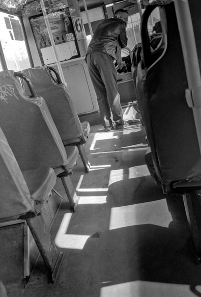 A monochrome photo shows the shabby interior of an old bus, which is flooded with sunlight. Shadows fall from the bus seats, creating a rhythm. A man stands by the driver's cabin.