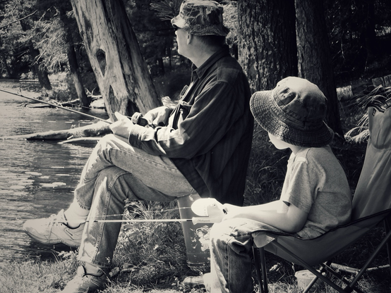 A young boy with his grandfather are seated along a lakeshore each holding fishing rods and each wearing bucket hats.