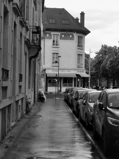 Rue de Nancy en noir et blanc un jour de pluie, une brasserie forme l'angle de la rue suivante