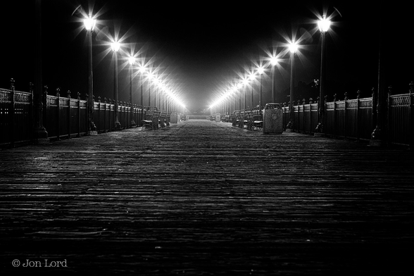 This Is A Very Dark And Grungy Black And White Street Photo, Without People, In Landscape Format. Stretching Upwards From The Base To A Little Over Half Way Up In The Frame Is A Surface Made Up Of Rough And Weathered Planks Of Wood Laid At Right Angles To The Camera. From Either Side Of The Image And Converging Inwards Towards The Middle Are Two Parallel Rows (Though They Appear To Converge) Of Metal Railings. Inset From Those Railings Are A Line Of Metal Street Lights, Fifteen To Twenty, That Again Appear To Converge Towards The Centre. The Tops Of Those Lights Have An Eight Point Star Glow, In Addition To Ever So Slightly Illuminating The Warm And Humid Air. To Add Some Municipal Ambiance To The Scene There Are Some Outward Facing Benches And A Few Wast Bins. 
The Location: Pier 7, San Francisco (2012)