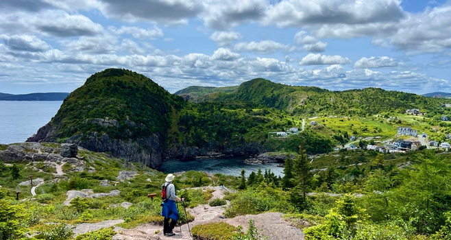 A rugged coastal landscape with a hiker in the foreground and the edge of a city in the background. The in the centre foreground hiker is facing away, wearing a tan brimmed hat, grey long shirt, red backpack and dark pants, with a blue jacket around the waist and holding hiking poles. The right side of the image shows closely spaced houses nestled around an ocean inlet, with a few houses scattered into the image centre. The rugged rocky and tree-lined coast is in partial sun cover under a blue sky with puffy white clouds.  The foreground shows winding rocky trails leading downhill with small green plants and shrubs. The overall coast colours are bright greens, with blue green water in the inlet. [Quidi Vidi (NL, Canada) June 2025]