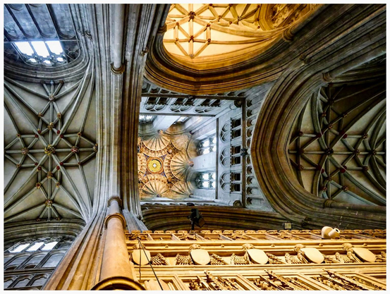 A view from below of the Canterbury cathedral's interior, showcasing intricate vaulted ceilings and decorative details. The image captures the towering columns and a beautifully crafted ceiling with ornate patterns and stained glass windows above.