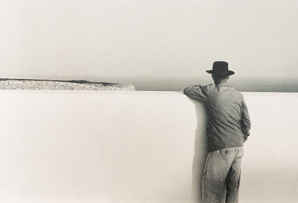 le bras appuyé sur un mur blanc lui arrivant aux épaules et partageant l’image horizontalement en deux, un homme de dos regarde ce qui est de l’autre côté, une falaise, la mer, l’horizon