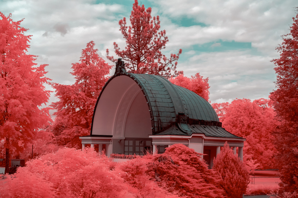 Das Bild zeigt einen Pavillon in einem Park mit Bäumen, die in infraroter Fotografie in verschiedenen Rottönen erscheinen.


Das Bild zeigt einen halbrunden Pavillon aus hellem Stein mit einem dunklen Dach, der inmitten eines Parks steht. Um den Pavillon herum befinden sich zahlreiche Bäume und Sträucher, die alle in einem intensiven, leuchtenden Rosarot erscheinen.  Dies deutet auf eine Bearbeitung des Bildes hin, wahrscheinlich mittels Infrarotfotografie, die die Pflanzenwelt in solchen Farben darstellt.  Der Himmel ist leicht bewölkt, in einem blassen Blau- und Weißton gehalten, was einen eher weichen und verträumten Kontrast zu den kräftigen Farben der Vegetation bildet. Die Gesamtstimmung des Bildes ist ruhig und friedlich, fast schon surreal durch die ungewöhnliche Farbgebung. Es wirkt harmonisch und idyllisch, ein ruhiger Ort zum Verweilen. Es sind keine Menschen oder Tiere auf dem Bild zu sehen. Die Architektur des Pavillons ist klassisch und elegant, was zur ruhigen und würdevollen Atmosphäre beiträgt.
