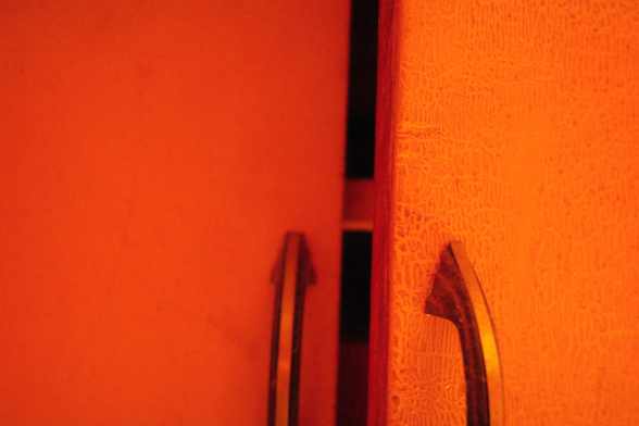 This photograph shows the gap between two cabinet doors, both painted or lit in intense orange. Each door has a dark metallic handle, with the right one attached to a textured, crackled surface. A narrow shadowed space is visible between the doors, revealing a faint glimpse of interior shelving inside.