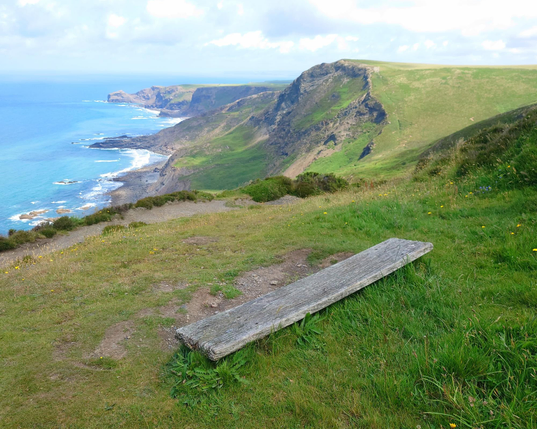 A seascape of blue ocean, slightly greying cumulus above, and headlands with green vegetation on the cliffs  and fields on top stretching off on the right hand side, with the foreground showing a simple weathered and low backless bench nearest the camera, which is angled towards sitting and taking in the view. beyond.