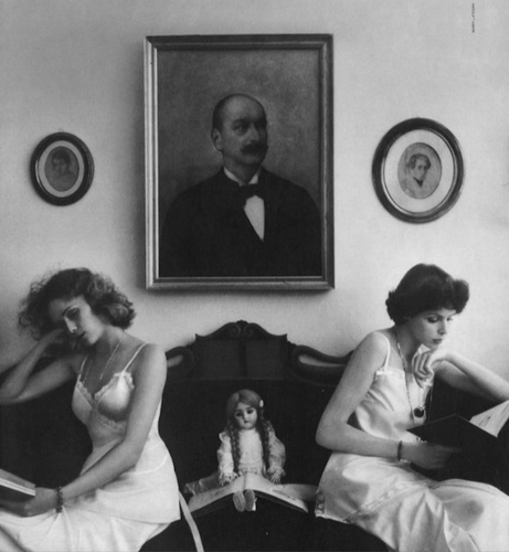 sur un canapé, deux jeunes femmes et une poupée, au dessus d’elles, le portrait du père moustachu dans son cadre en bois