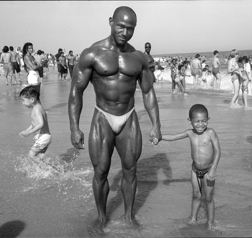 sur la plage, les pieds dans l’eau, un petit garçon souriant donne la main à son père culturiste aux muscles puissants luisant au soleil