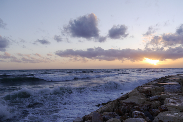 Eine heitere Küstenszene bei Sonnenuntergang mit rollenden Wellen, die an eine felsige Küste prallen, unter einem farbenfrohen Himmel mit vereinzelten Wolken.