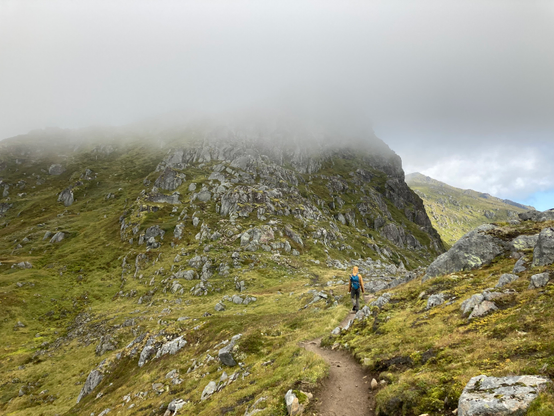Zu sehen ist ein schmaler Wanderweg, der sich über einen grünen Berg schlängelt. Es sind vereinzelte nackte Felsen zu sehen. Die Wolken hängen sehr tief. In einiger Entfernung ist eine Person auf dem Weg zu sehen.