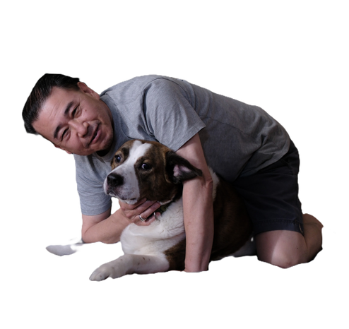 A man in a gray shirt on his knees holding his white and brown dog. The background is removed.