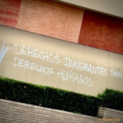 Graffiti sprayed on the side of a concrete building reads Derechos immigrantes son derechos humanos. 
Above the tag, windows have been covered with protective plywood.
