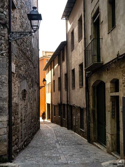 Color photo of a person walking down a narrow stone-paved street passing from a shadowed area to a sunlit area. 