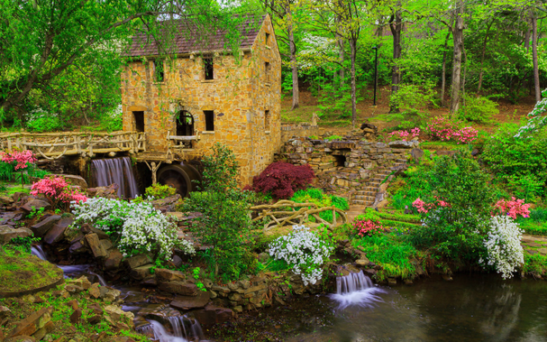 A horizontal image of a picturesque scene of a rustic stone mill nestled amidst a lush, vibrant landscape. A cascading waterfall adds to the serene ambiance, and the surrounding greenery is bursting with life. The mill is a quaint, two-story building constructed from weathered stone. Its roof is a blend of dark, aged shingles and natural wood, giving it a charming, rustic appearance. A large, wooden water wheel is attached to the side of the mill, suggesting its historical purpose. The mill is beautifully framed by a variety of plants and flowers. Bright pink and white azaleas dot the landscape, their colorful blooms adding a cheerful touch. Lush green trees and bushes provide a backdrop of natural beauty, with their leaves creating a canopy of shade. A wooden bridge gracefully spans a small stream that flows from the waterfall, inviting visitors to explore the surroundings. A cascading waterfall takes center stage in the image. The water flows over rocks and into a small pond.