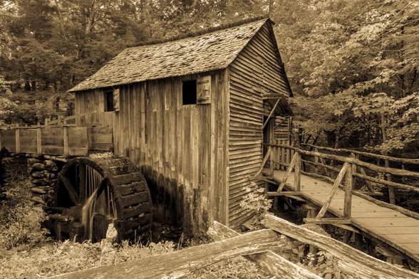 A sepia-toned, horizontal image of the historic John P. Cable Grist Mill in Cades Cove, Tennessee. The mill, a rustic, two-story wooden structure, stands nestled among dense trees. To the left of the building, a large wooden waterwheel, dark with age and dampness, is partially visible, indicating the mill's function. A small amount of water is visible at the bottom of the wheel. The wooden flume that would feed water to the wheel is visible behind it, connecting to the mill building. On the right side of the mill, a wooden walkway with railings extends from the building, leading into the treeline. In the immediate foreground, a rustic split-rail fence runs diagonally across the bottom of the frame.