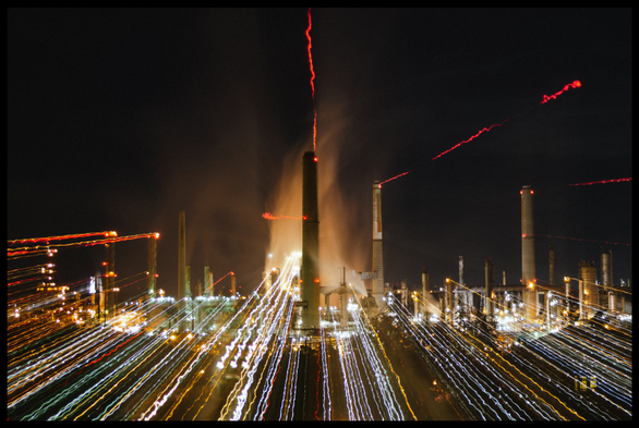 Some colorful eye candy: an oil refinery, brightly lit at night, shining against dark skies. The yellow, red, orange, and green spotlights of the refinery explode outward with shaky lines, moving at high speed towards the camera. In the center: a large smokestack surrounded by orange hazy steam that rises slowly into the sky, almost flame-like.