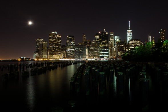 A nighttime skyline of a city with illuminated buildings, a bright moon in the sky, and old pier pilings in the water in the foreground.