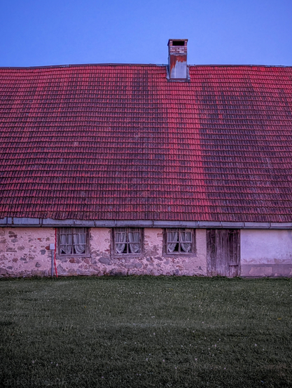 The front of a rustic, aged farmhouse, under a twilight sky. In front of the building, a stretch of green grass extends across the bottom of the frame.

The most dominant feature is the expansive, steeply pitched roof covered in reddish-brown tiles. The tiles show signs of weathering, with some areas darker or lighter. A single chimney, made of brick and topped with a metal flue, protrudes from the ridge of the roof on the right side. 

Below the roof, the building's facade is a mix of textures and materials. There are three windows, evenly spaced, each with light-colored curtains drawn inside. To the far right, there's a wooden door. 