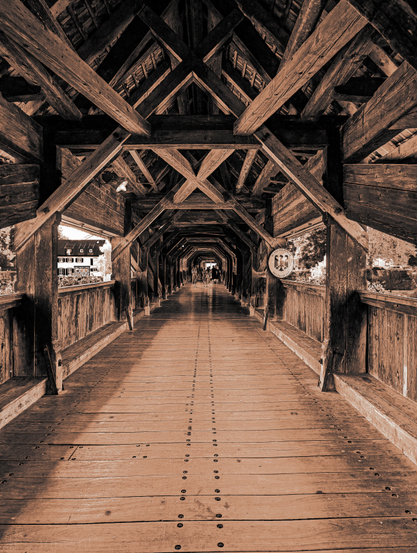 A sepia-toned, interior shot of a long, wooden covered bridge. The perspective looks straight down the length of the bridge, emphasizing its tunnel-like structure and the repetitive wooden beams of its ceiling. The bridge's construction is prominently featured, with exposed wooden trusses forming an intricate overhead pattern. 

The light at the end of the tunnel effect is prominent, drawing the eye towards the vanishing point where a few figures can be seen in the distance. Through the openings on the left side, a building with white walls and a dark roof, is partially visible. The overall tone of the image is warm and historic due to the sepia filter, enhancing the wooden textures and giving the scene a timeless quality.

The Holzbrücke Bad Säckingen (German name) or Säckingerbrücke (Swiss name) is a bridge over the Rhine. It connects the German city of Bad Säckingen with the village of Stein in Switzerland. The covered bridge spans 203.7 metres over the Hochrhein and is the longest roofed wooden bridge in Europe. 