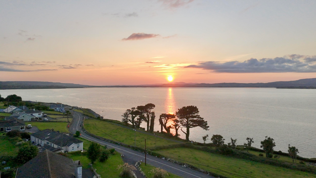 View of the sun setting over Dungarvan Bay from a drone flying about 50m above Helvick looking north west on June 15th. 

The sun is just below a narrow bank of cloud extending to the north just above the horizon. 

A column of reflected orange light spans the bay from just below the sun to a small copse of yew trees on the shore.  A couple of houses can be seen on the left in the foreground with two large fields reaching down to the shore beyond a road that winds from the left of the picture to the lower right roughly following the south coast of the bay. 