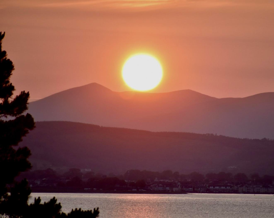 View of the sun setting behind  Knockmealdown mountain over Dungarvan Bay viewed from Helvick looking north west on June 15th. 

The picture is a long zoom shot centered on the sun with the sun’s disc  just touching the distant mountain before it sets behind it. The exposure and long zoom highlights the distance between the mountains in the far background that are distinctly lighter in tone due to atmospheric haze than the foreground hills. The coast line around Dungarvan below the hills is closer and therefore darker still and the distance framing is completed by the silhouette of the edge of a tree in the foreground on the left that is darker still. 
