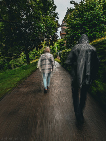 A slightly blurred wet, dark asphalt path lined with lush green trees and bushes. Two figures are walking away from the viewer in the rain. The figure on the left is more clearly defined. The figure on the right is much more blurred and appears almost like a dark, indistinct silhouette. Both people are wearing a poncho.