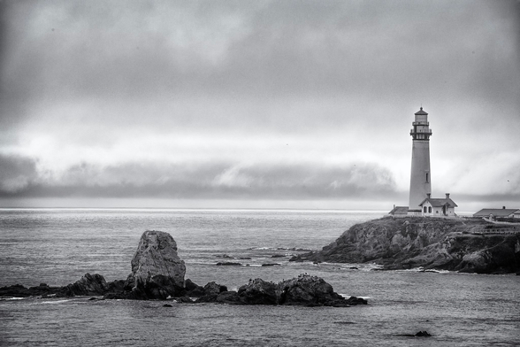 Black and white photo of Pigeon Point lighthouse on a foggy morning. In addition to the lighthouse, we see a rocky outcropping offshore.