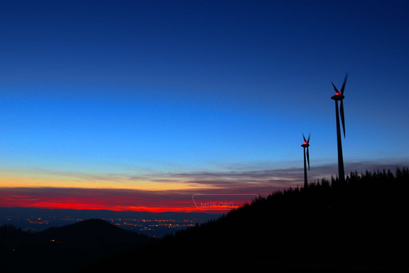 Eine Landschaftsaufnahme im Vordergrund sind ein schwarzer Berg mit zwei Windkraftwerken. Im Hintergrund ist ein Tal mit beleuchteten Gebäuden und einem Himmel in der blauen Stunde mit roten Wolken. / A landscape shot in the foreground is a black mountain with two wind turbines. In the background is a valley with illuminated buildings and a sky in the blue hour with red clouds.