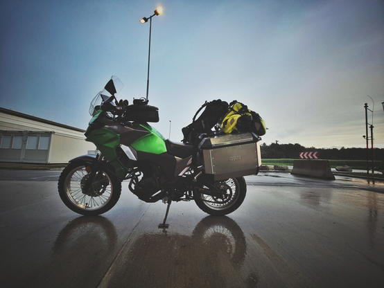 A low-angle, eye-level full shot of a green and grey Kawasaki Versys X-300 motorcycle, laden with luggage, parked on a wet asphalt surface which reflects the bottom part of the motorcycle. The motorcycle is oriented to the left, with a Givi Alaska aluminum pannier visible on the right side and a soft bag on the passenger seat. The sky is cloudy with a hint of brightness on the horizon. A street light pole stands to the left of the motorcycle, and some white buildings are visible in the background on the left. To the right, there are concrete barriers and a red and white chevron sign.