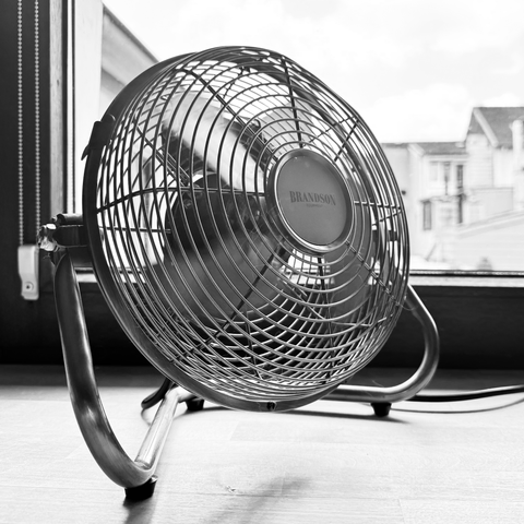 Schwarz-weiß-Aufnahme eines Ventilators vor einem geschlossenen Fenster. Im Hintergrund sind Häuser zu sehen.