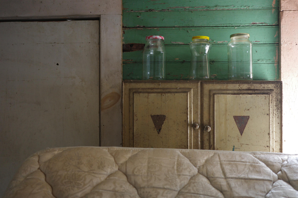 This photograph shows a worn indoor scene with a cream-colored quilted cushion or mattress in the foreground. Behind it sits a distressed yellow cabinet with triangular symbols on its doors, topped by three empty glass jars with colored lids (red, yellow, and beige) arranged against a wall of peeling green wooden planks. To the left of the cabinet is an aged white door with smudges, stains, and a small hole, framed in rough, slightly darker wood.