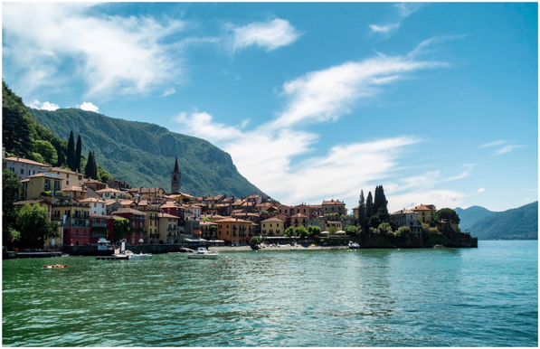 A picturesque lakeside village with colorful buildings along the shore, surrounded by mountains and greenery. The scene features calm water, a few boats, and a clear blue sky with wispy clouds.